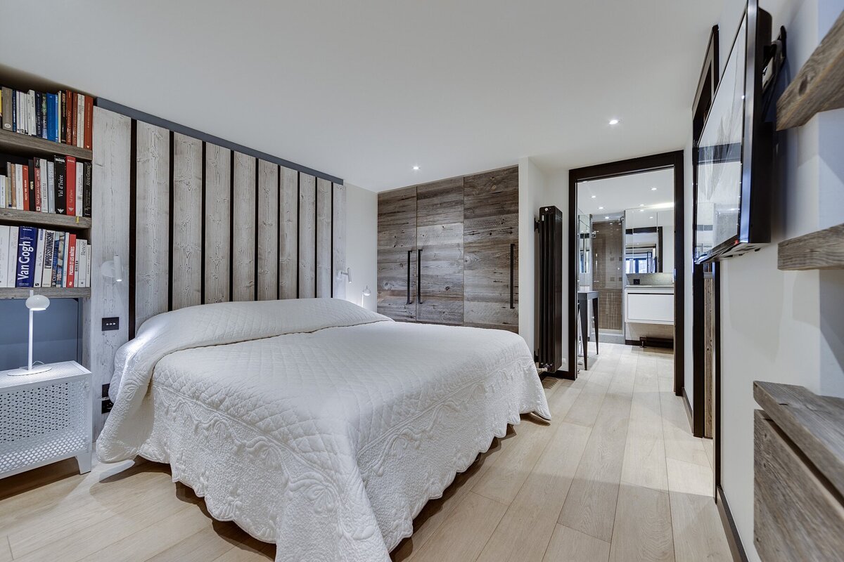 A stylish bedroom with a large white bed, distressed wood paneling, a bookshelf, and a dark wooden wardrobe. An ensuite bathroom is visible beyond.
