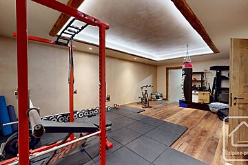 A spacious home gym with a red power rack, punching bag, exercise bike, and weights on wood and rubber flooring, featuring ambient ceiling lights and exposed beams.