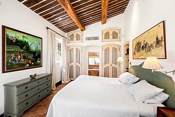 A rustic bedroom with exposed wooden ceiling beams, white walls, built-in wardrobes, a dresser, and paintings.