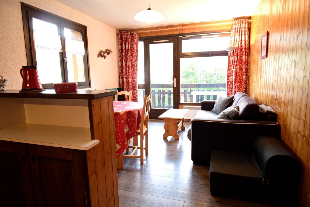 Cozy cabin-style living room with wooden walls, a dark sofa, a dining table with red tablecloth, and balcony doors.