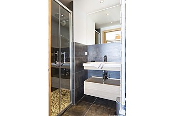 A modern bathroom features a glass shower with a pebble floor, a floating vanity with a sink and large mirror, all set against dark gray tiles.