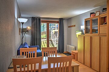 A bright, compact living/dining area with wooden furniture, a blue sofa, and a glass door overlooking outdoor stairs. Grey walls and wooden floors.