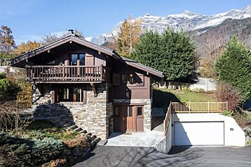 A cozy stone and wood alpine chalet with a garage, set against majestic snow-capped mountains on a sunny day.