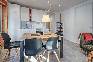 A modern kitchen and dining area featuring white cabinets, a wooden table with black chairs, and grey tiled flooring. An armchair with a red blanket sits nearby.