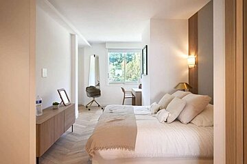A bright, modern bedroom with a large bed, neutral colors, and a desk area by a window, featuring light wood accents and a clean design.
