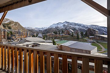 Balcony view of a mountain ski resort with wooden buildings, green fields, and snowy peaks under a clear sky.
