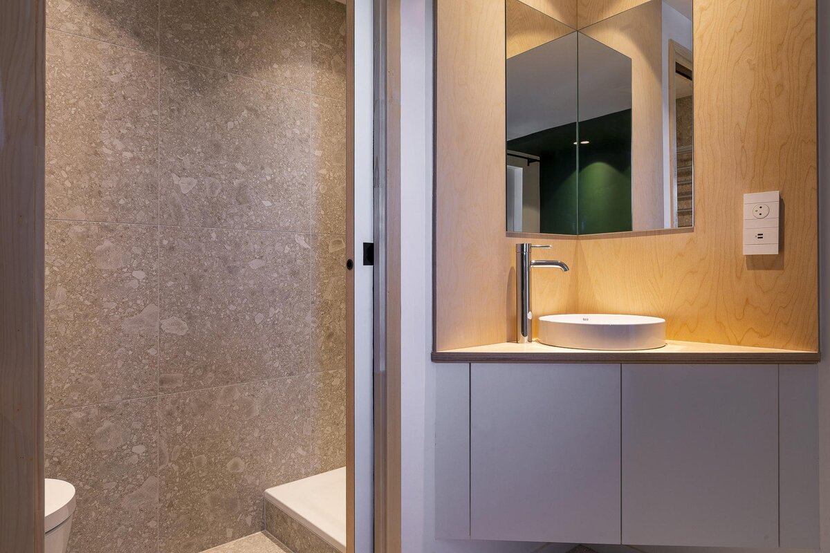 Contemporary bathroom with plywood vanity, vessel sink, mirror, and lighting. Grey tiled shower area visible nearby.