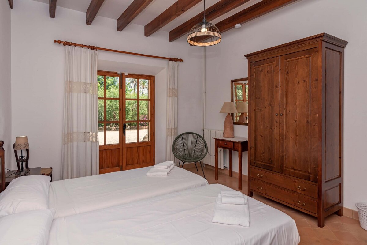 A cozy bedroom with two beds, exposed wooden beams, and a door opening to a lush garden. Features a rustic wooden armoire and a unique round chair.