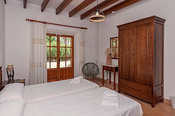 A cozy bedroom with two beds, exposed wooden beams, and a door opening to a lush garden. Features a rustic wooden armoire and a unique round chair.
