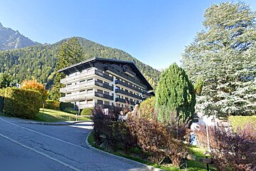 A multi-story building with balconies, set against forested mountains under a clear sky. A road curves in the foreground, surrounded by trees and bushes.