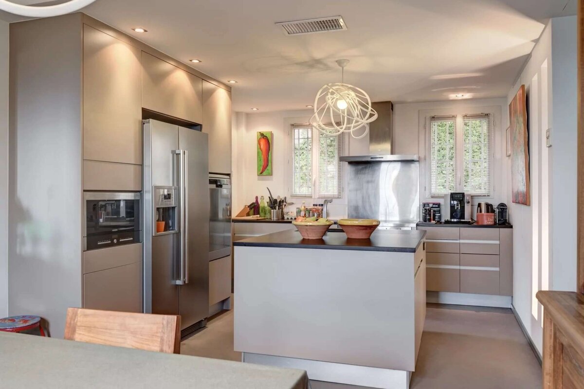 A modern kitchen with neutral cabinets, stainless steel appliances, a central island, and a unique white pendant light. Windows with blinds provide natural light.