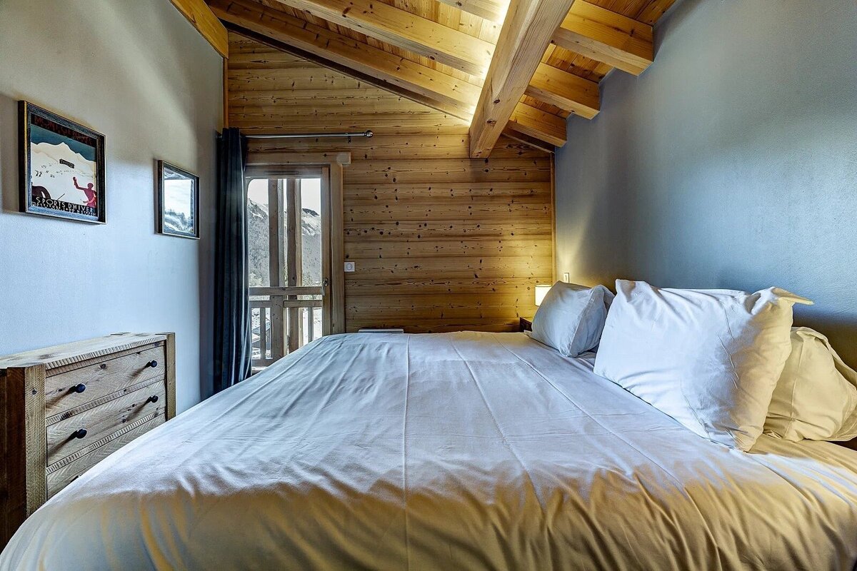 Cozy chalet bedroom with wood paneling, white-linened bed, ski-themed art, and a balcony offering snowy mountain views.