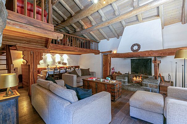 Cozy, rustic chalet living room features exposed wood beams, a stone fireplace, comfy sofas, a dining area, and a loft overhead.