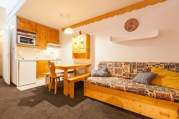 Cozy studio apartment with a rustic wooden kitchen, dining table with benches, and a patterned sofa bed on a dark carpet.