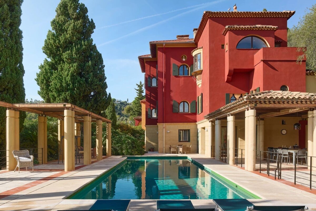 A grand Mediterranean villa with red walls and a swimming pool flanked by columned pergolas, set against tall green trees.