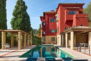 A large red villa with a swimming pool, flanked by covered patios and tall green cypress trees.