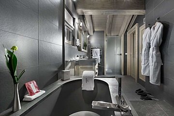 A luxurious, modern bathroom features dark grey tiles, a sleek black tub, double sinks, a glass shower, and exposed wooden beams. Robes and slippers are ready.
