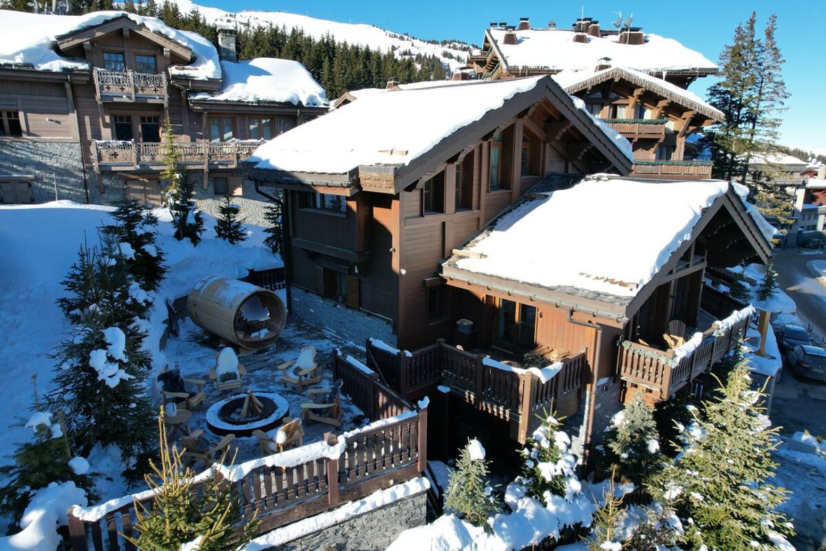 Snowy alpine village with wooden chalets, an outdoor fire pit with chairs, and a barrel sauna, all under a clear blue sky.
