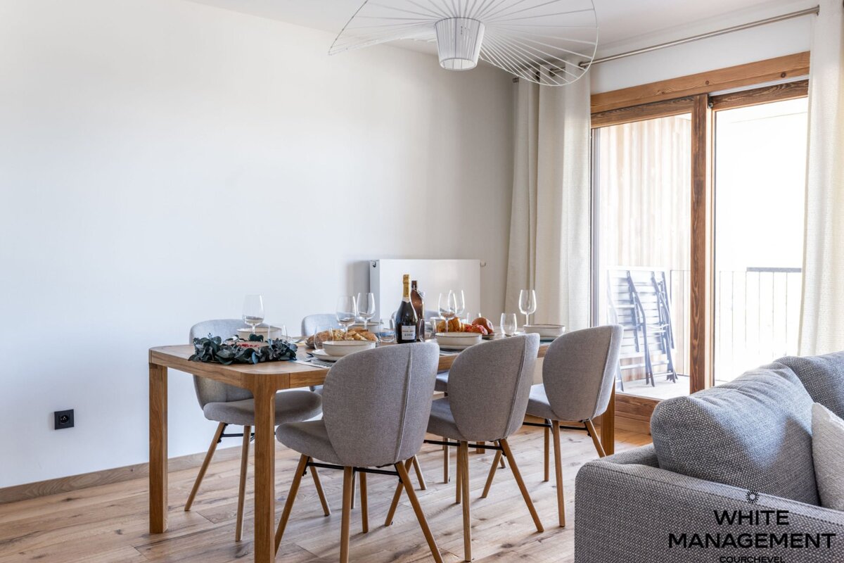Modern dining area with a light wooden table and grey chairs, set for dinner. A contemporary chandelier hangs overhead, and a balcony is visible.