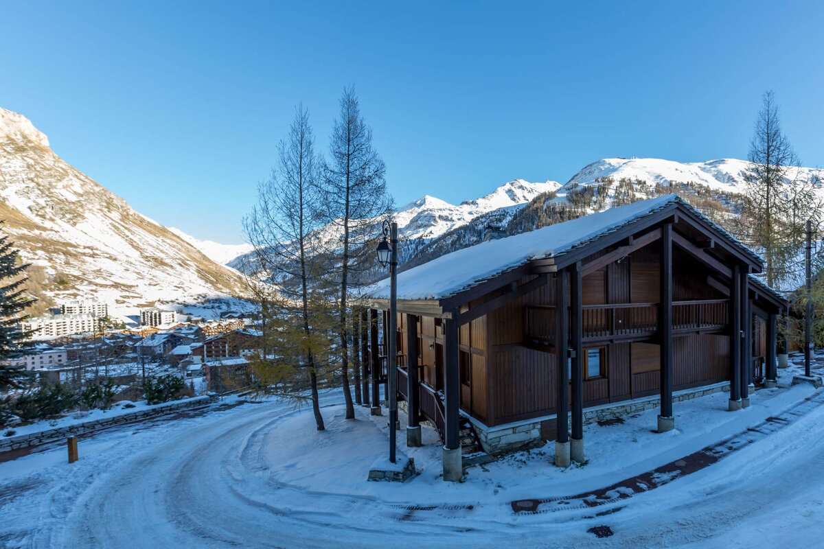 A snow-dusted wooden chalet stands beside a winding snowy road, overlooking a mountain village. Snow-capped peaks rise under a clear blue sky in this winter scene.