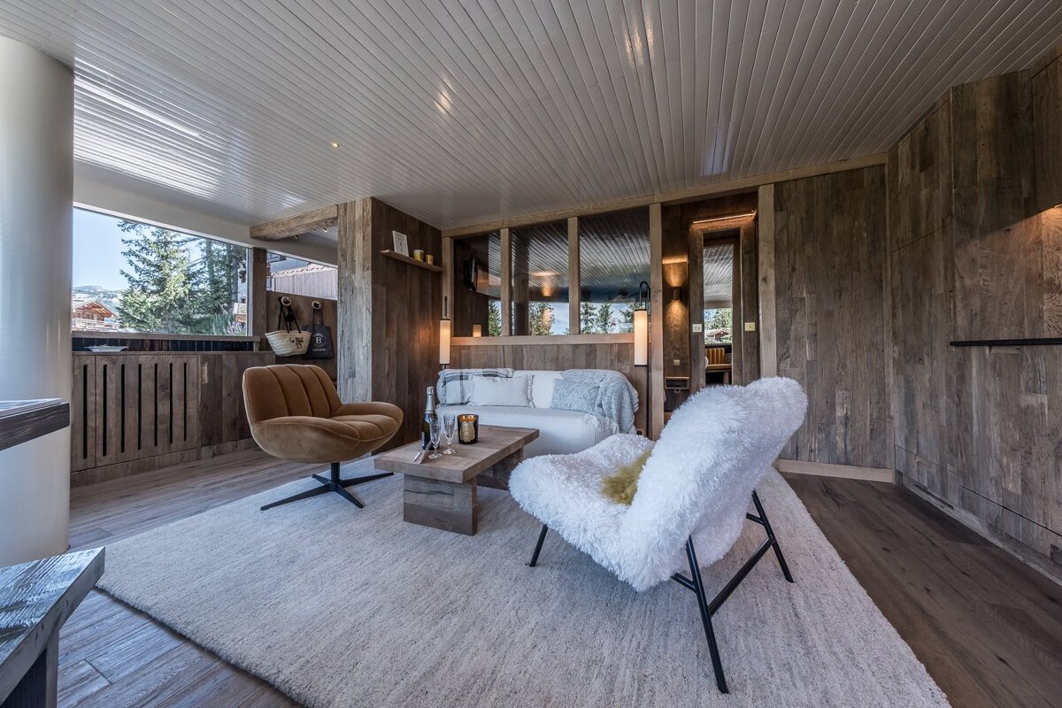 Cozy chalet living room with rustic wood walls, a white fuzzy armchair, a brown swivel chair, and a large area rug.
