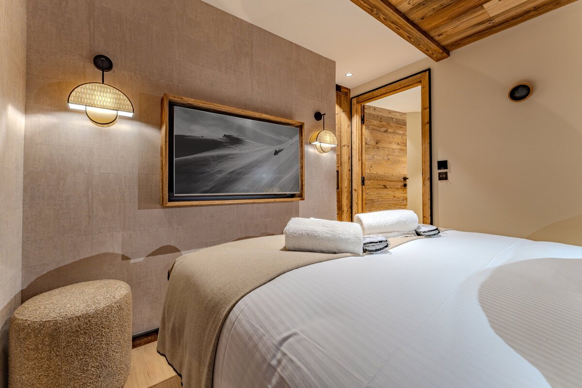 A cozy, neutral-toned bedroom with a bed, white linens, a TV showing a snowy landscape, and rustic wood accents. Woven wall sconces provide soft light.
