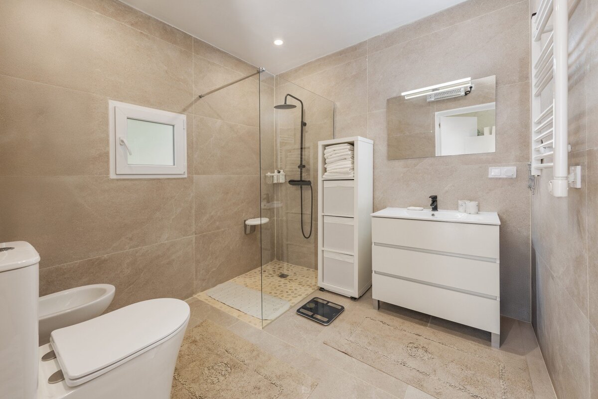A modern beige bathroom with a white toilet, bidet, glass shower, white vanity, mirror, storage unit, and towel warmer.