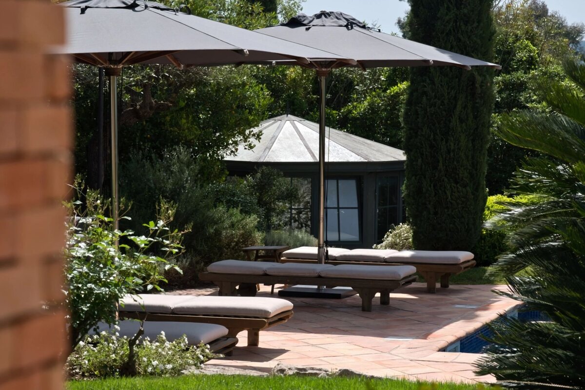 Wooden loungers with cushions sit under large grey umbrellas on a tiled patio next to a pool, surrounded by lush gardens.
