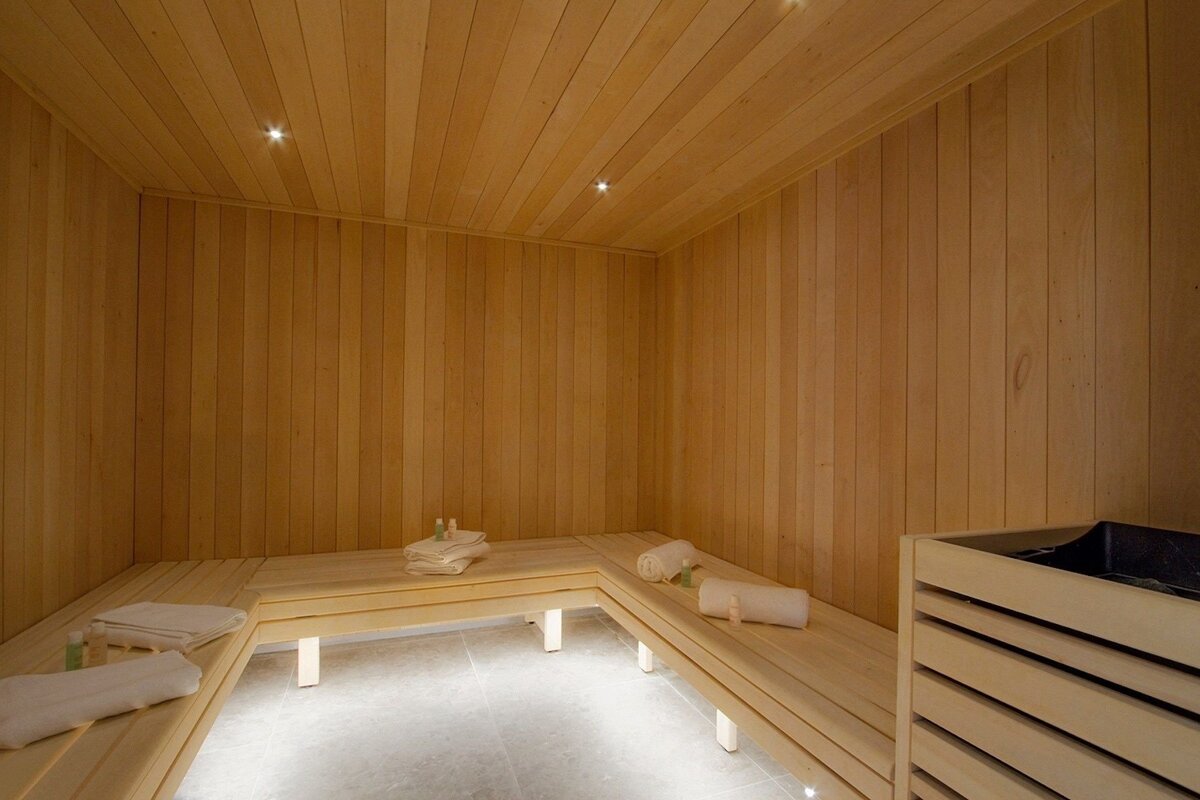An empty, modern wooden sauna featuring light-colored wood paneling, benches lined with towels, and soft under-bench lighting.