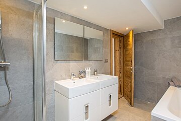 Modern grey-tiled bathroom featuring a double sink vanity, mirrors, a glass shower, and a rustic wooden door.