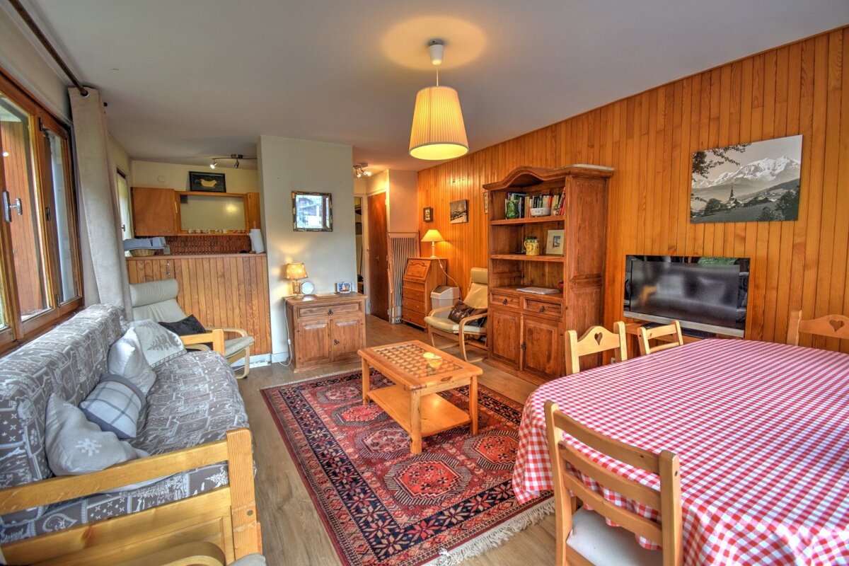 Cozy, wood-paneled living/dining room with rustic wooden furniture, including a sofa, shelving unit, and dining table with a checkered cloth.