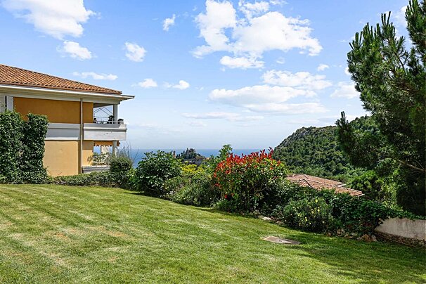 A yellow house with a lawn and garden, offering views of the blue sea and forested mountains under a partly cloudy sky.