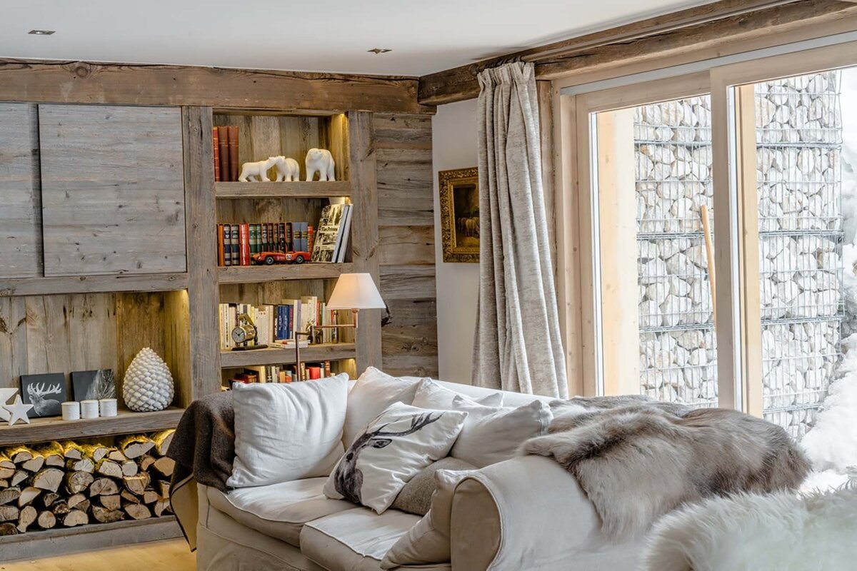 A cozy, rustic living room with wood-paneled walls, built-in bookshelves, a comfy sofa, and a window overlooking a stacked stone wall.