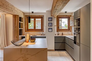 Modern kitchen with wood beams, a marble island, and gray cabinets. Chalet-style windows and mountain-themed artwork.