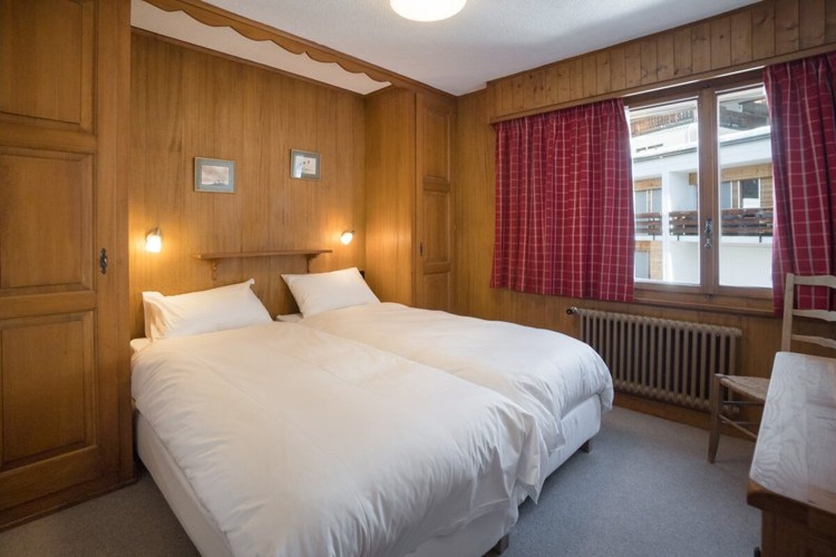 Rustic wood-paneled bedroom with twin beds pushed together, white linens, and a window with red checkered curtains.