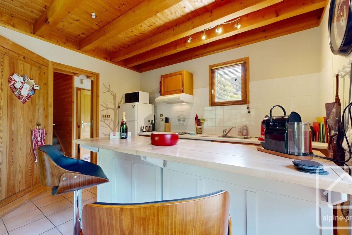 A rustic-modern kitchen with wooden ceiling beams, a light-colored breakfast bar, a window, and essential appliances. Champagne bottle visible.