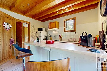 A rustic-modern kitchen with wooden ceiling beams, a light-colored breakfast bar, a window, and essential appliances. Champagne bottle visible.