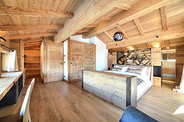 A luxurious chalet-style bedroom features extensive light-toned wood on walls, floor, and exposed ceiling beams, with a large bed and a modern pendant light.