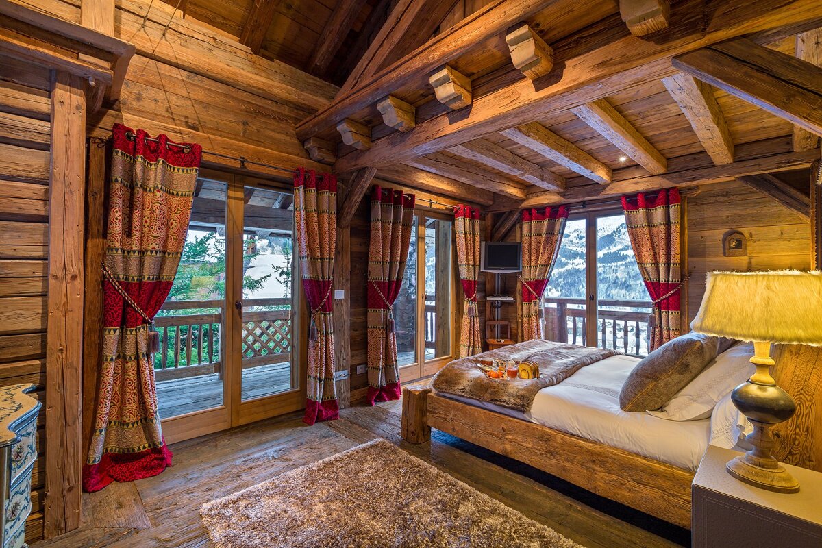 Cozy wooden chalet bedroom with a large bed, vibrant red curtains, and stunning snowy mountain views from the balcony doors.