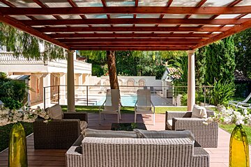 A luxurious outdoor patio with wicker furniture under a pergola, overlooking a pool and a large house with columns, surrounded by lush greenery.