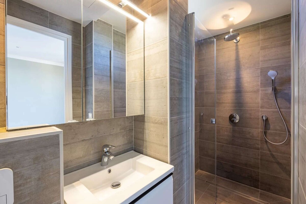 A contemporary bathroom with a white sink, large mirror with LED light, and a glass-enclosed shower, all with grey tiles.
