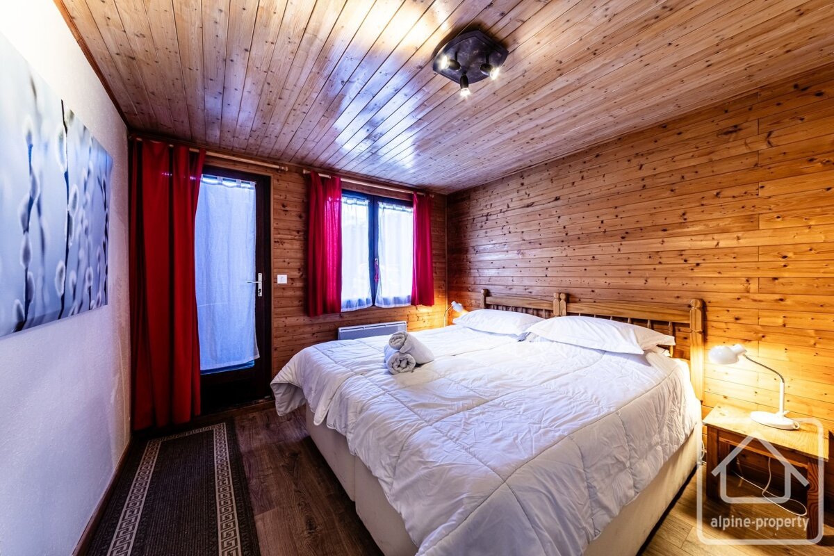 A cozy, rustic bedroom featuring extensive wood paneling, a large bed with white linens, red curtains, and warm lighting, plus blue/white artwork.