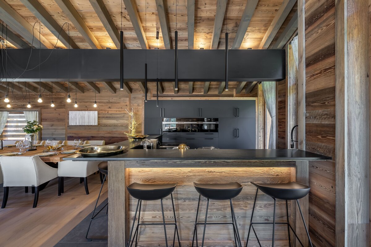 A luxurious modern chalet kitchen and dining area with rich wood, dark sleek finishes, and sophisticated lighting.