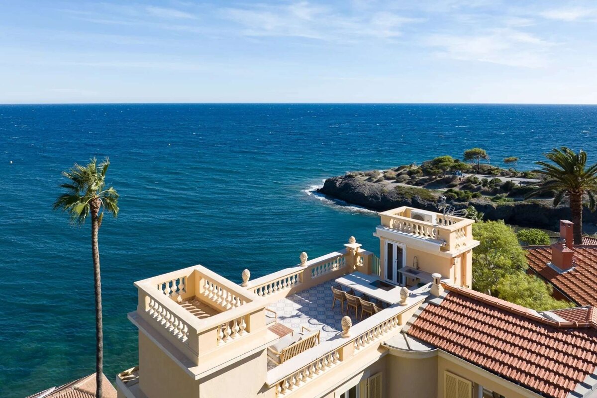 An aerial view shows a luxurious coastal villa with a rooftop terrace overlooking the vibrant blue sea, a rocky peninsula, and a tall palm tree.