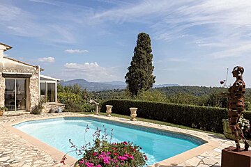 A stone villa with a sparkling pool and modern sculpture, offering panoramic views of mountains and lush greenery under a clear blue sky.