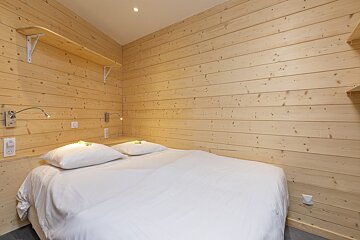 A cozy bedroom with light wood paneling on all walls, featuring a large bed with crisp white bedding, and two wall-mounted reading lights.