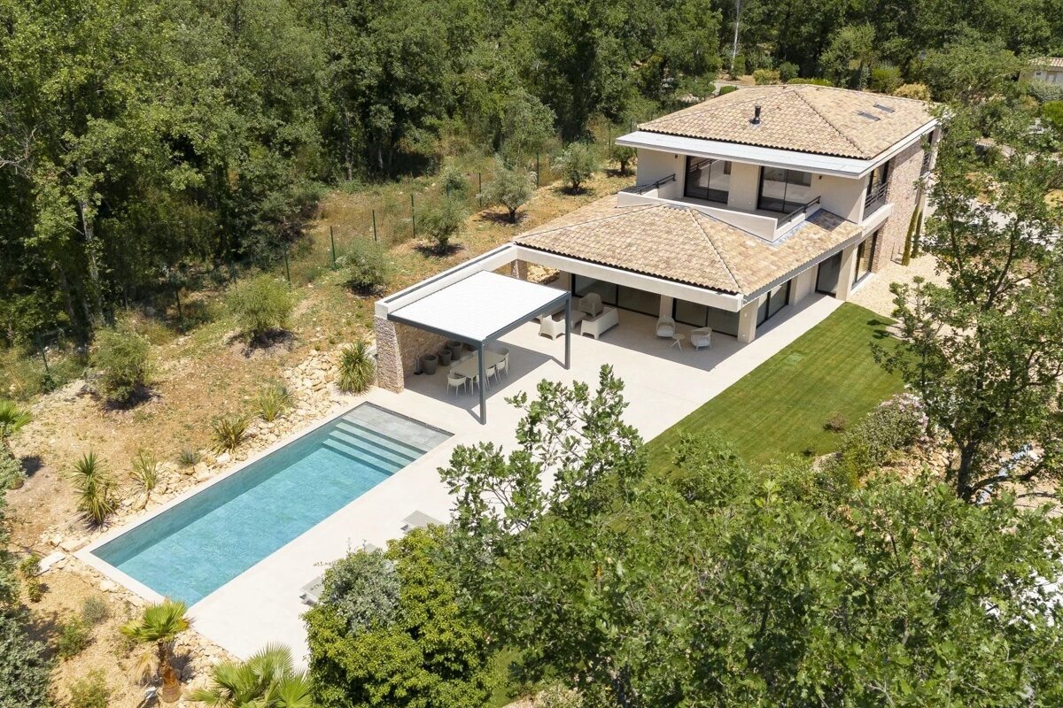 An aerial view of a modern two-story villa with a tiled roof, featuring a swimming pool, patio, and lush green surroundings.