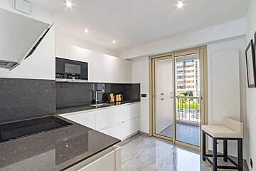 A sleek, modern kitchen with white cabinets, dark gray countertops, and built-in appliances. A sliding glass door opens to a balcony overlooking city buildings.