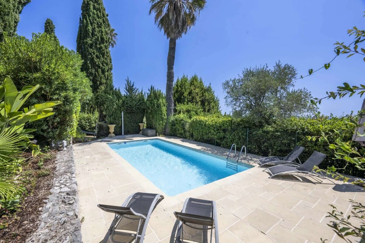 A sunny outdoor rectangular pool with clear blue water, surrounded by beige patio tiles, four sun loungers, and lush green trees including cypresses and a palm.