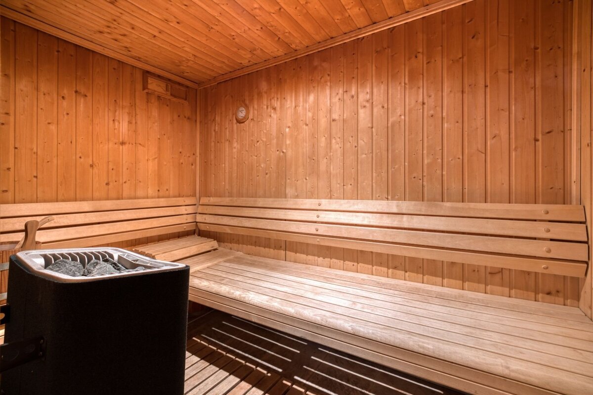 A bright, empty wooden sauna room with slatted benches and a black sauna heater with stones, illuminated by warm light.